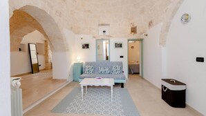 A cozy living room with a sofa, a small table, and a patterned rug. There is a doorway leading to another room, and a small cabinet is visible in the corner. - Trullo Paleo (Ceglie Messapica)