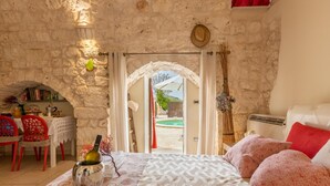 Um quarto aconchegante com parede de pedra, cama com roupa de cama estampada, área de jantar com cadeiras vermelhas e vista através de uma porta arqueada.