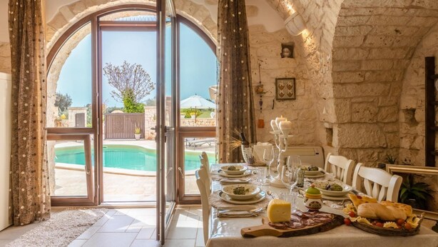 Uma área de refeições com uma mesa posta para refeição, piscina visível através de uma porta aberta e paredes de pedra.