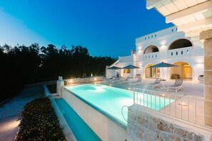 Un área junto a la piscina con sillones y sombrillas, un muro de piedra y un edificio con ventanales arqueados.