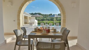 Un espace de restauration en plein air avec une table dressée pour quatre personnes, entourée de chaises en osier, donnant sur une piscine et une végétation luxuriante.
