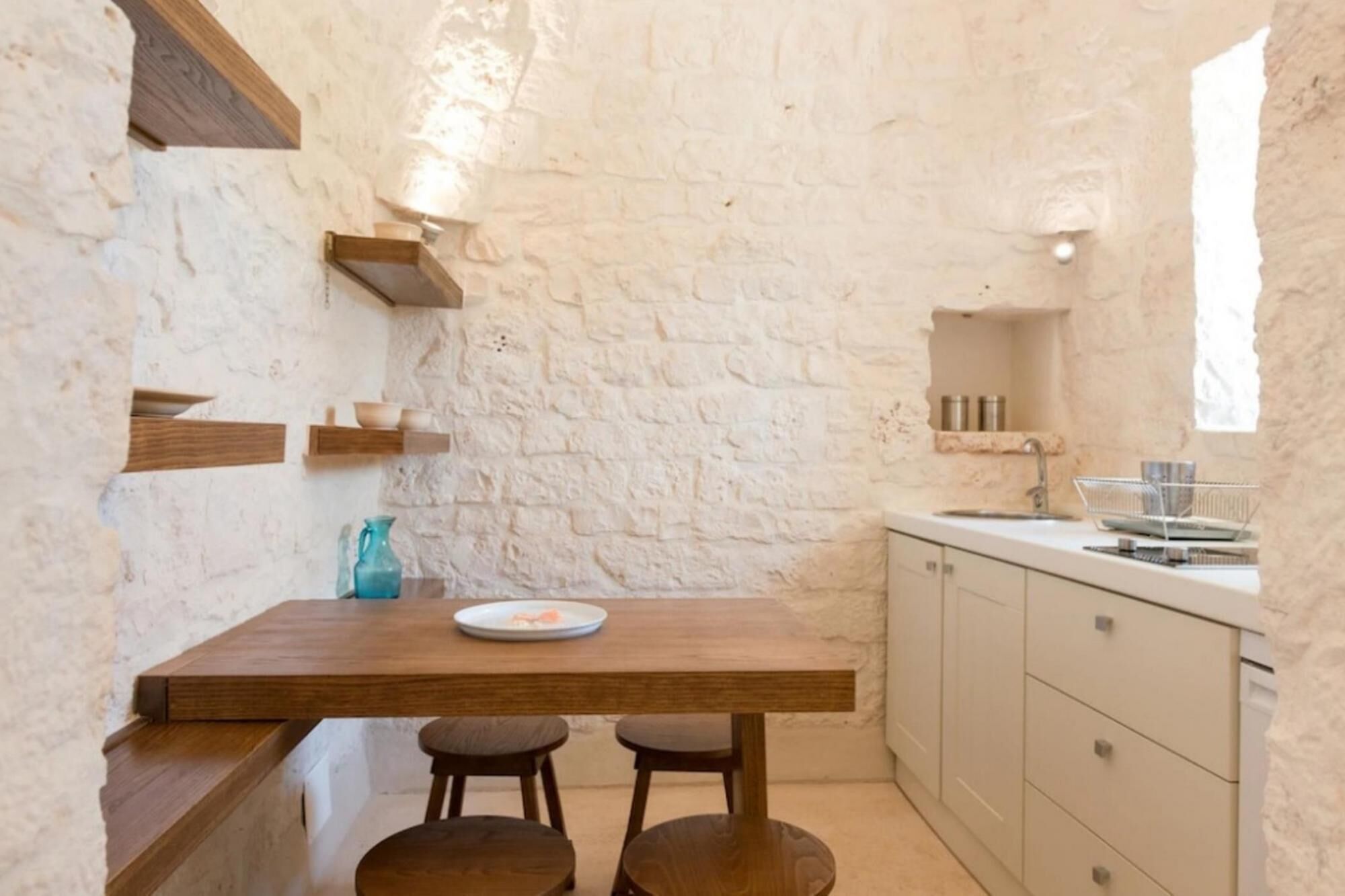 A compact kitchen with a wooden table, two stools, a white sink, and a window.
