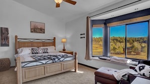 Quarto com cama de madeira, mesa de cabeceira, luminária, janela com vista para árvores e ventilador de teto.
