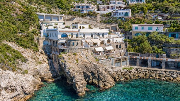 Un edificio costero con fachada de piedra, varios balcones y aguas azules cristalinas al fondo.