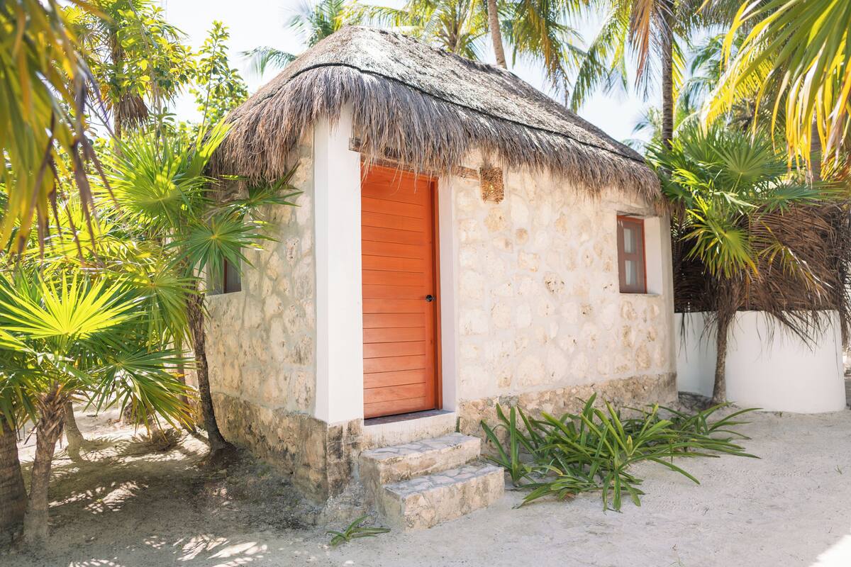 A small, thatched-roof hut with a single wooden door and steps leading up to it, surrounded by palm trees and sandy ground.