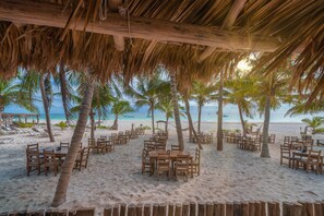 Un área de comedor frente a la playa, con mesas y sillas de madera, palmeras y vista al océano.