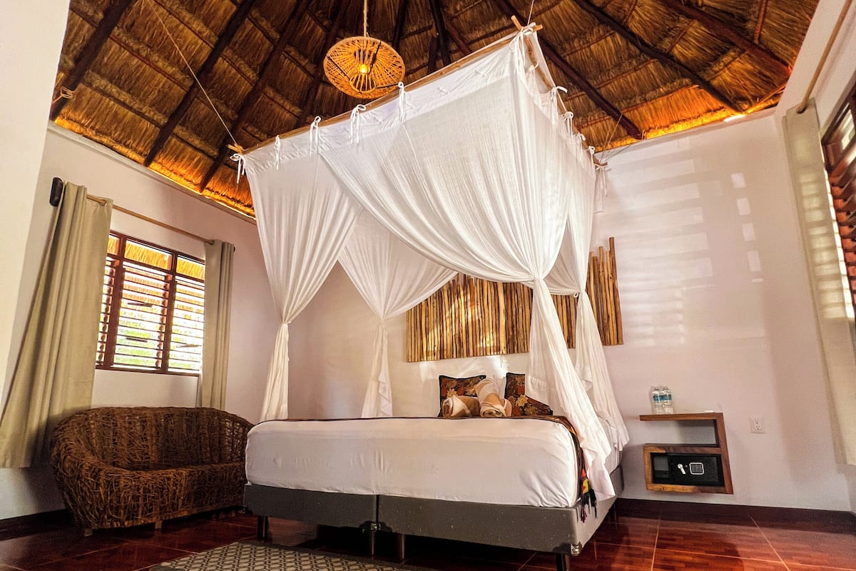 A room with a bed under a canopy, a sofa, a small table with a water bottle, and a wall-mounted safe.