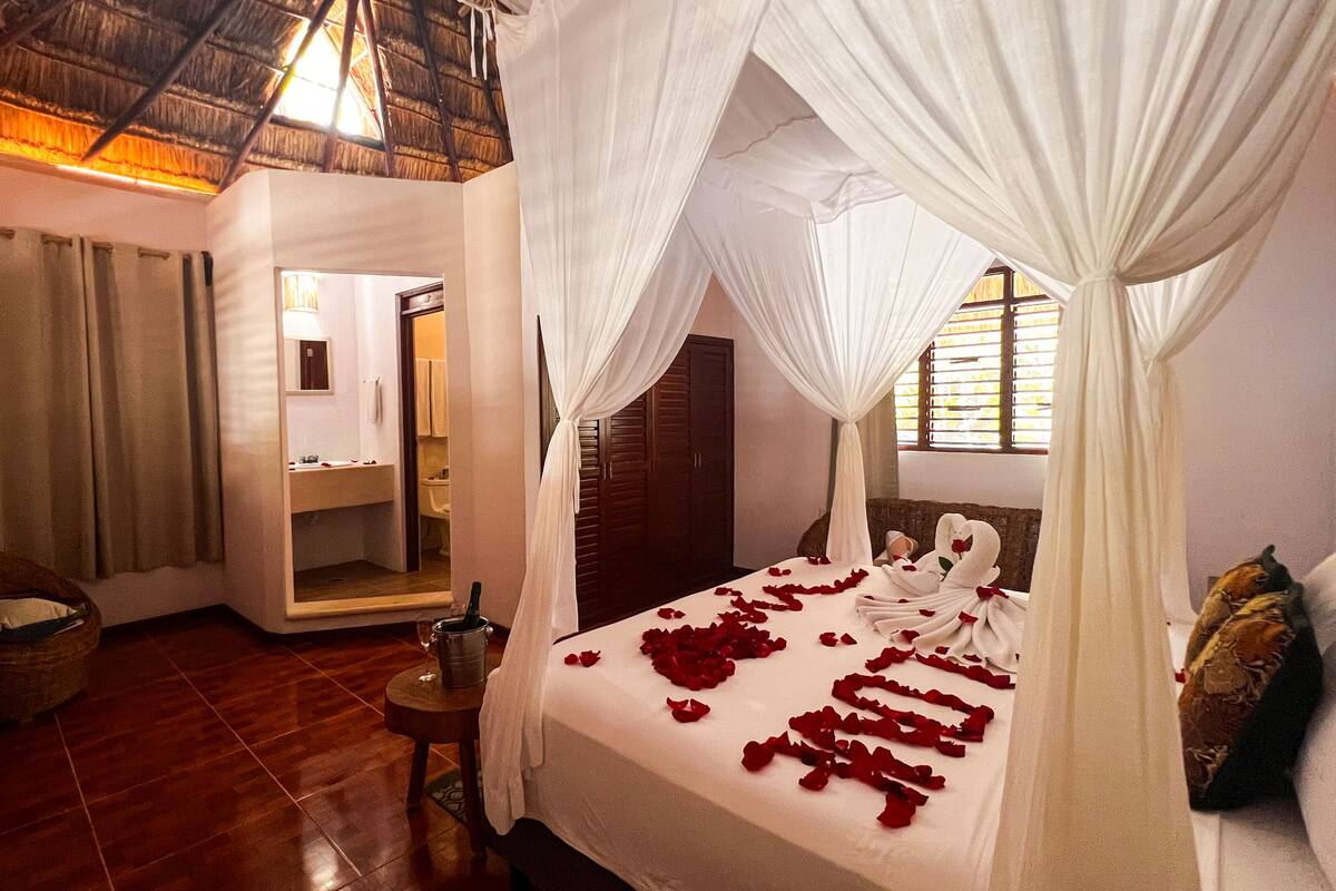 A room with a bed adorned with rose petals, a mirror, a wooden floor, and a thatched roof.