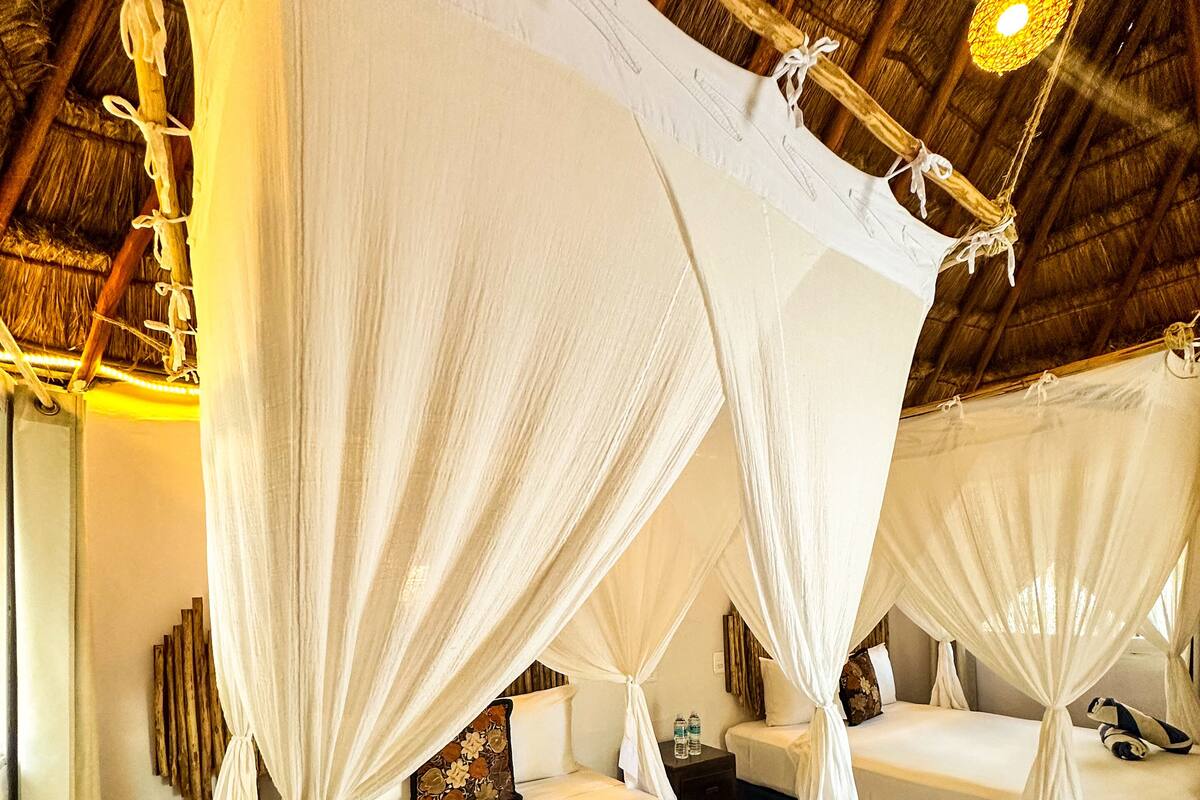 A room with four beds, each with a canopy bedspread and a pillow. The room has a thatched roof and a wooden floor.