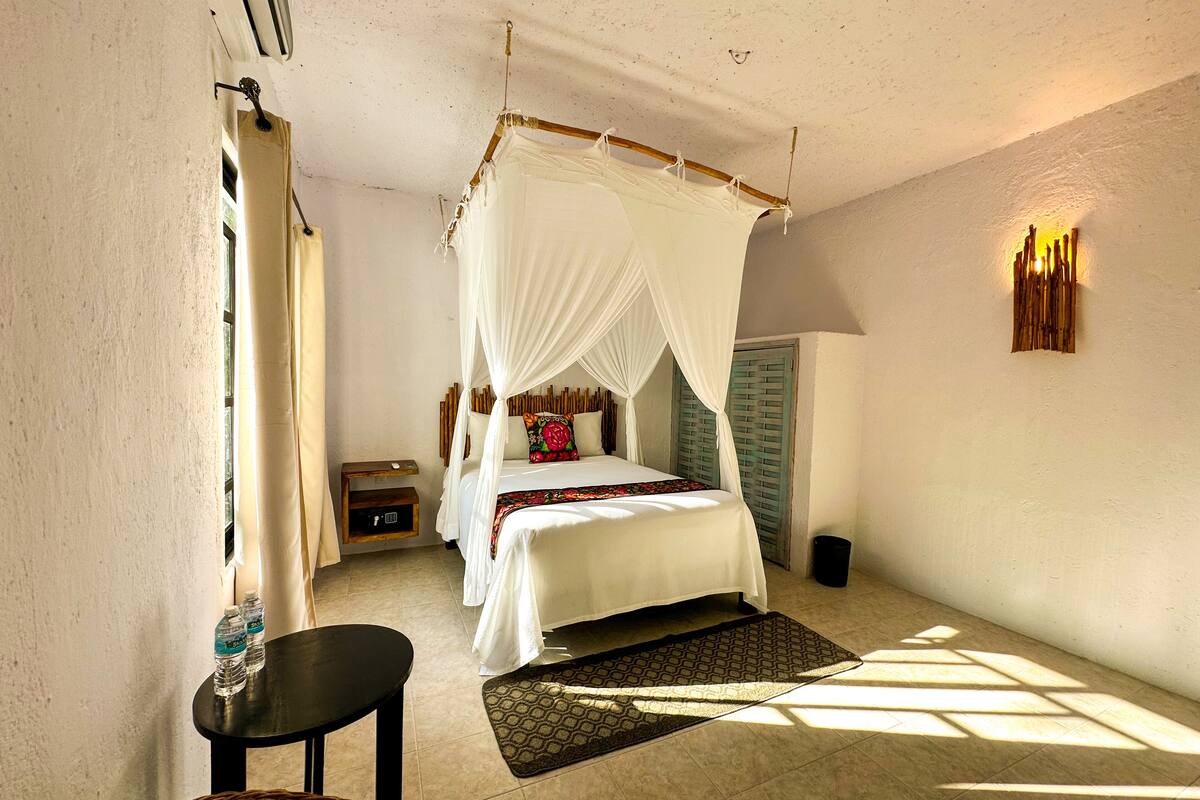 A bedroom with a canopy bed, a bedside table with a lamp, a small wooden nightstand, and a woven chair.