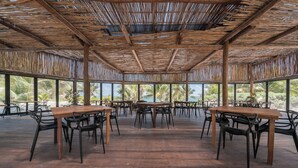 An open-air dining area with wooden tables and chairs, large windows, and a thatched roof. - Alito Tulum Hotel (Tulum)