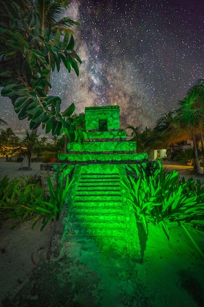 Una estructura piramidal iluminada en verde, rodeada de vegetación tropical bajo un cielo estrellado.