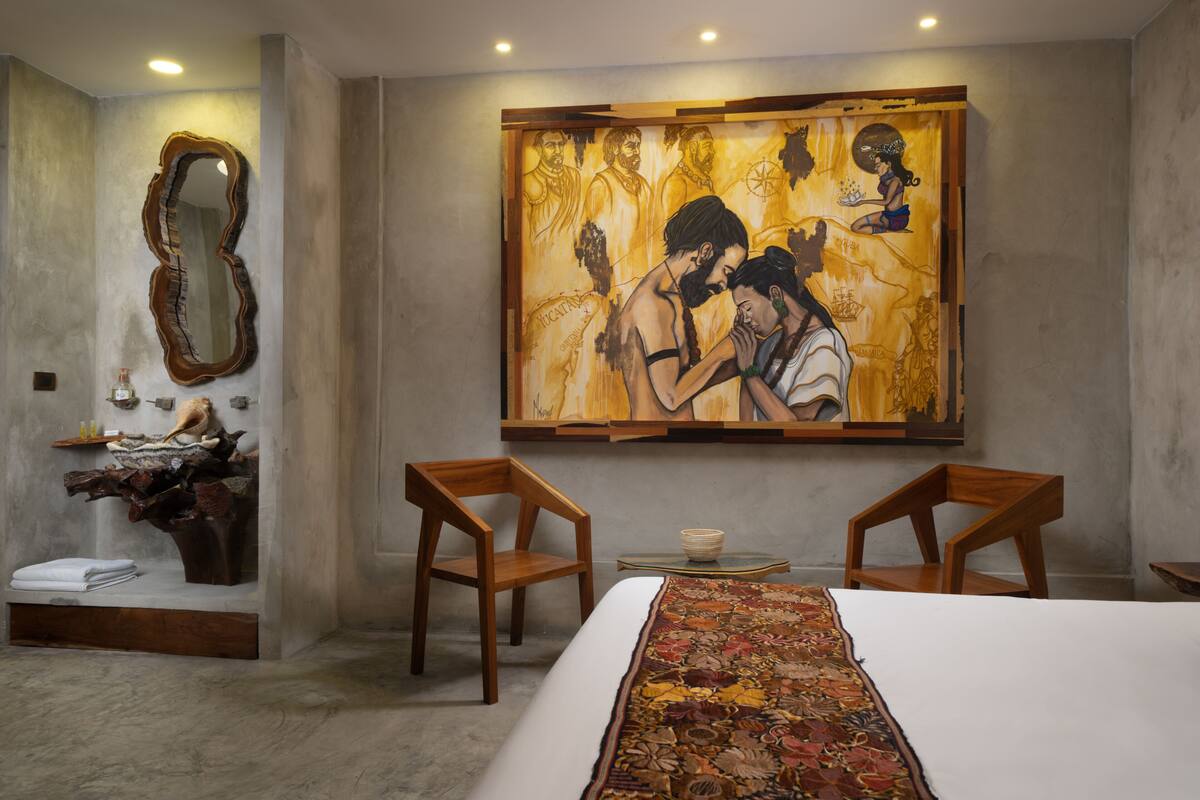 A hotel room with a large painting, a wooden chair, a small table, and a bed with a patterned bedspread.