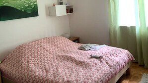 Une chambre à coucher avec un lit, un luminaire suspendu, une étagère fixée au mur, un tableau et une fenêtre avec des rideaux.