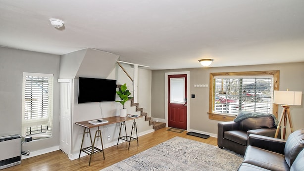 A living room with a wooden floor, a red door, a TV mounted on the wall, a brown leather couch, and a small table with stools.