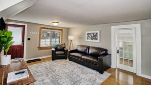 A living room with a brown leather couch, a wooden coffee table, a patterned rug, a red door, a window with blinds, and a framed picture on the wall.