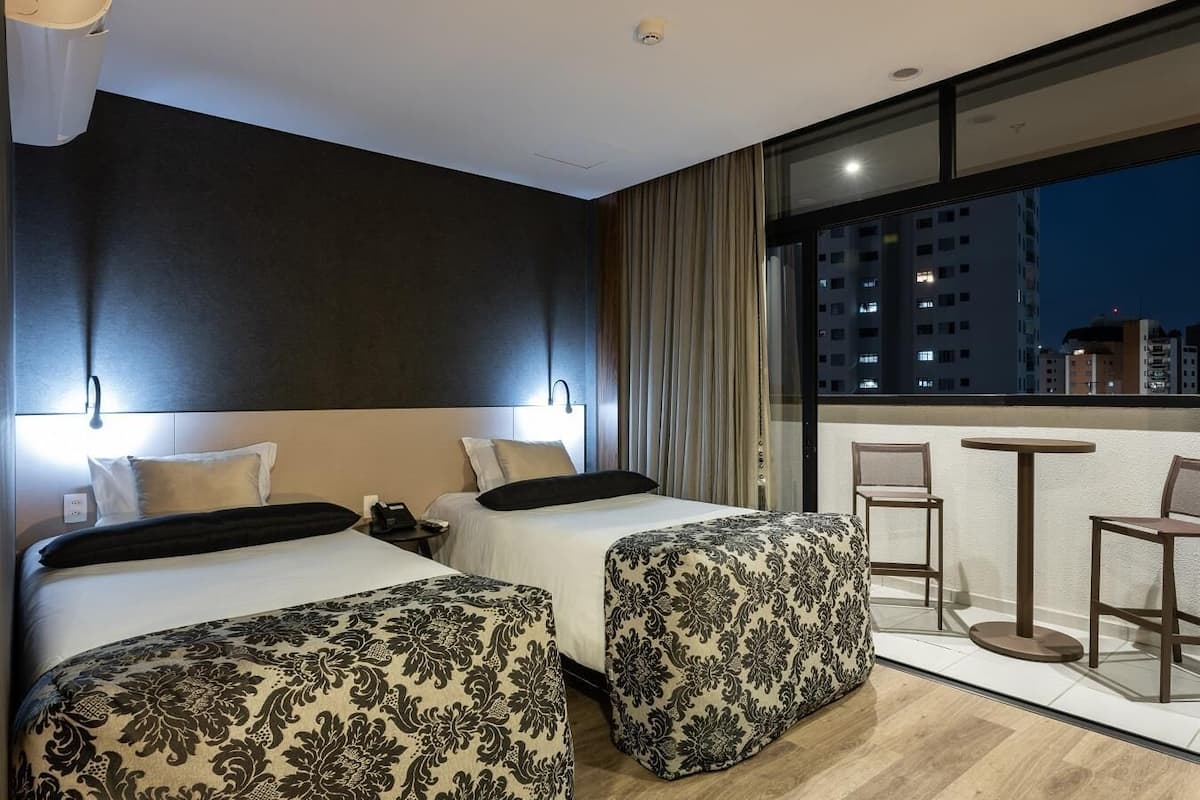 Quarto de hotel com duas camas, uma varanda com cadeiras e vista para a cidade à noite.