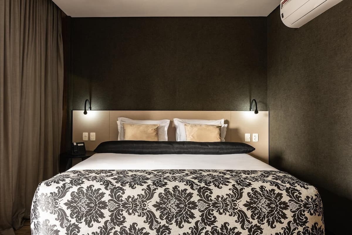 Quarto de hotel com uma cama grande, duas luminárias de cabeceira, um telefone e ar condicionado fixado na parede.