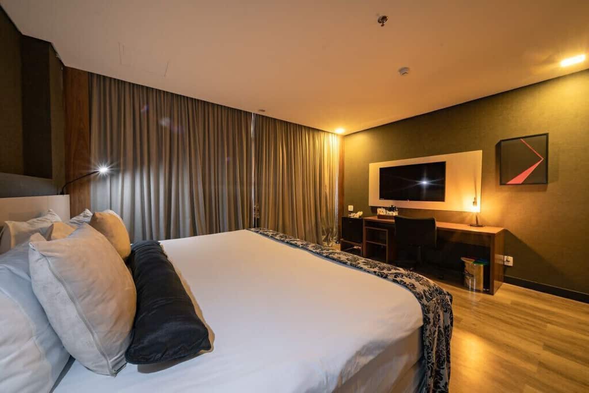 Quarto de hotel com uma cama, escrivaninha, cadeira, televisão e cortina.