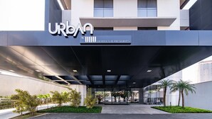 Un edificio moderno con un acceso cubierto, señalización de 'URBAN by UNU Hotels & Resorts', y un área ajardinada con árboles y arbustos pequeños.