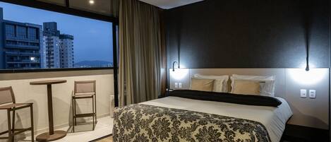 A hotel room with a large bed, a balcony with two chairs, and a city view.