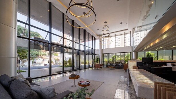 Un moderno vestíbulo de hotel con grandes ventanales, una zona de estar con sofás, una mesa de centro y una zona de bar con televisor.