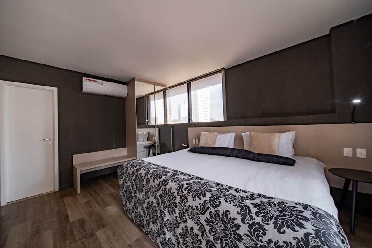 Quarto de hotel moderno com uma cama grande, um banco, uma mesinha e vista da cidade através de grandes janelas.