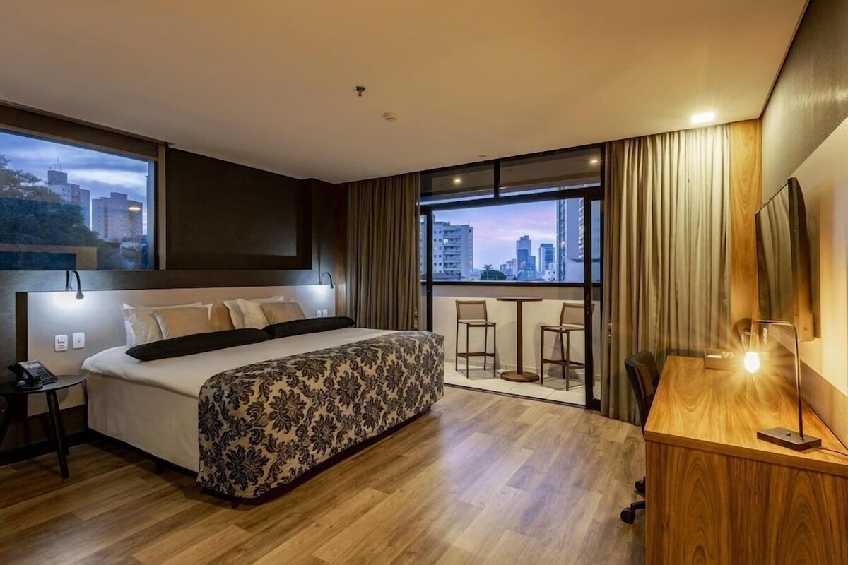 A modern hotel room with a large bed, a desk, and a balcony with a city view.
