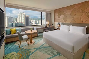 Ein Hotelzimmer mit einem großen Bett, einem Sofa, einem Schreibtisch und Blick auf die Stadtlandschaft.
