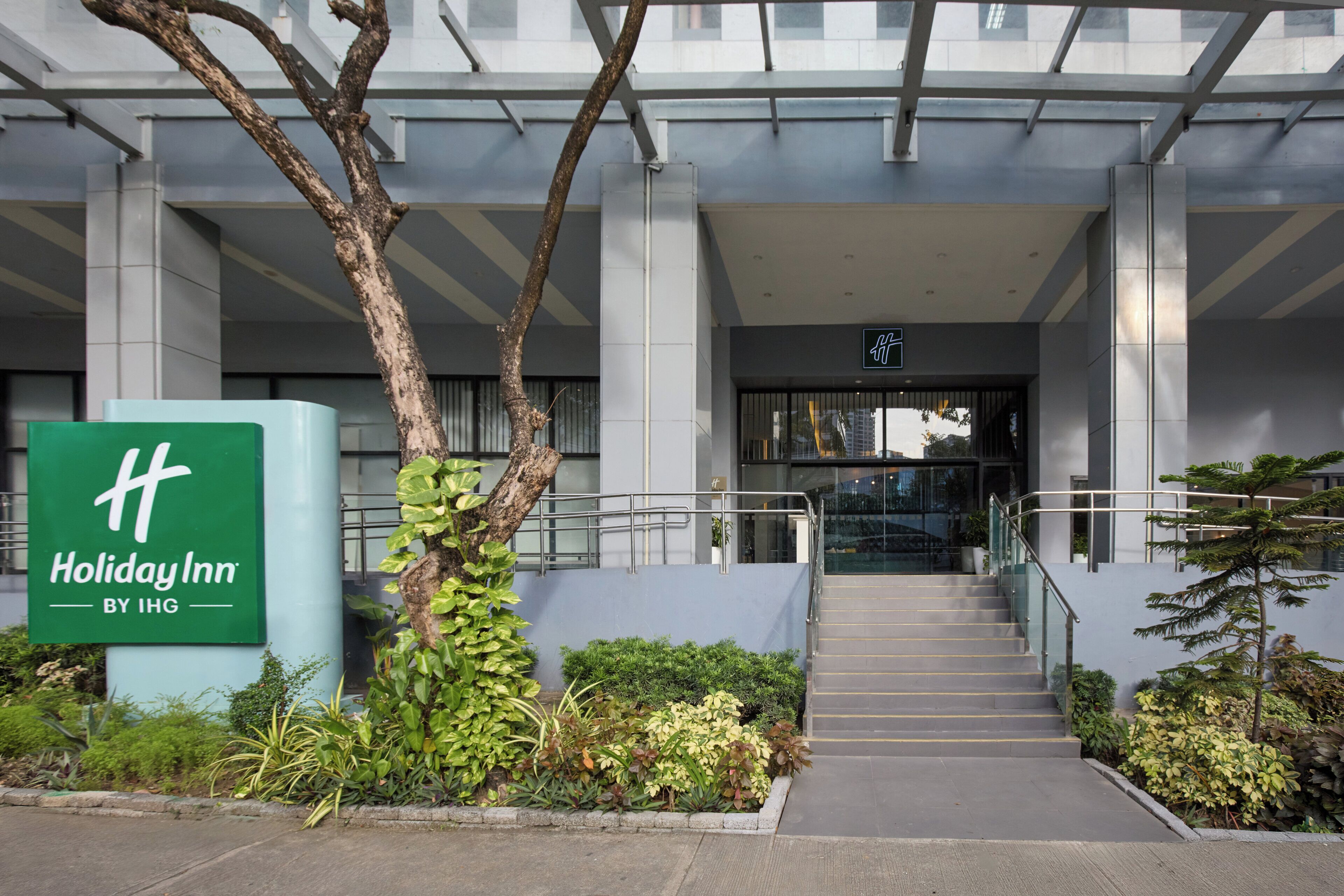 A Holiday Inn by IHG entrance with a staircase leading to the lobby, surrounded by greenery and modern architecture.
