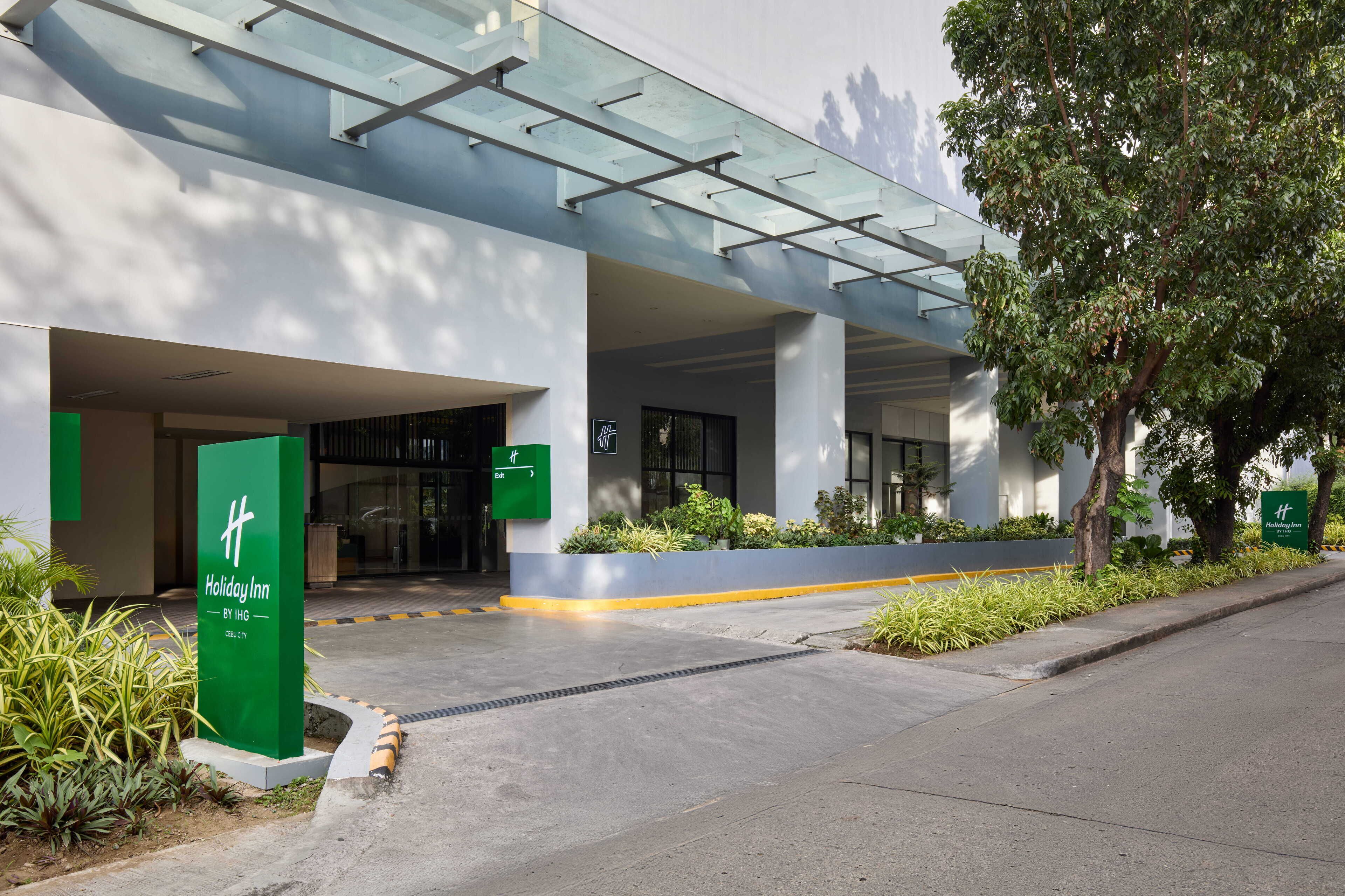 A modern hotel entrance with a glass canopy, green signage, and a landscaped pathway.