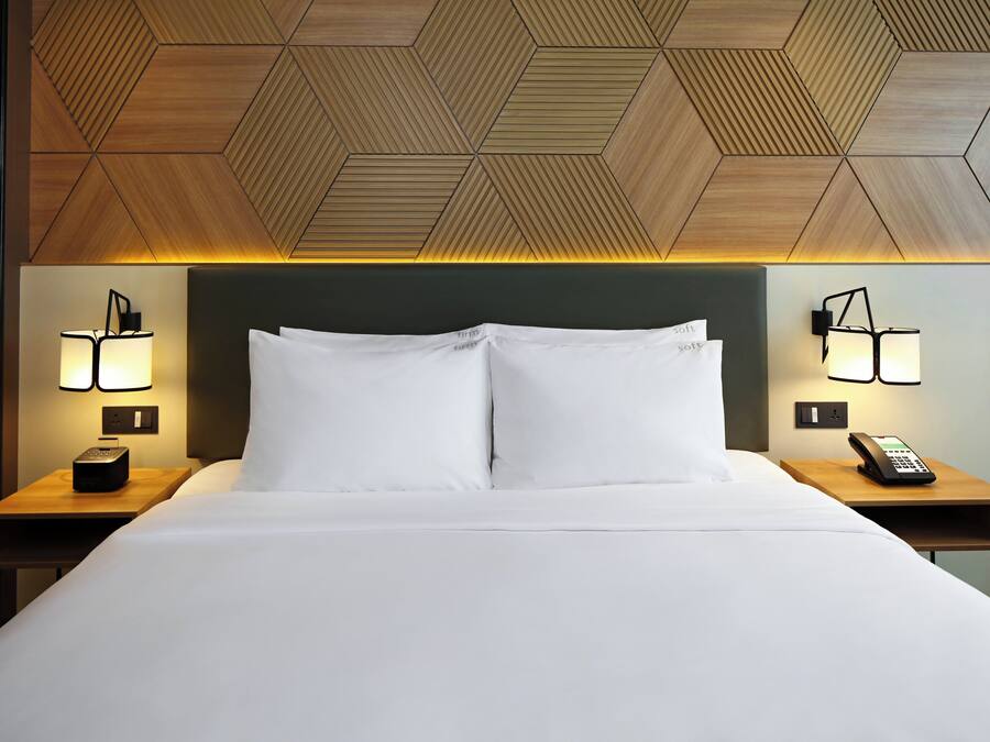 Une chambre d’hôtel avec un grand lit, deux tables de chevet avec des lampes, un téléphone et un plafond orné de motifs géométriques.