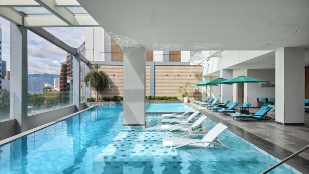 An indoor swimming pool with clear blue water, surrounded by lounge chairs and umbrellas, with a cityscape view through large windows. - Holiday Inn Cebu City by IHG (Cebu City)
