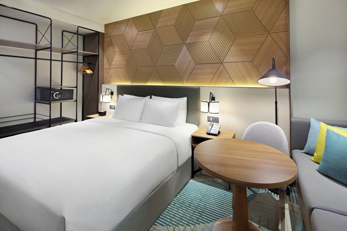 Une chambre d’hôtel moderne avec un grand lit, un canapé, une petite table ronde, un bureau avec une lampe et un mur orné d’un motif géométrique.