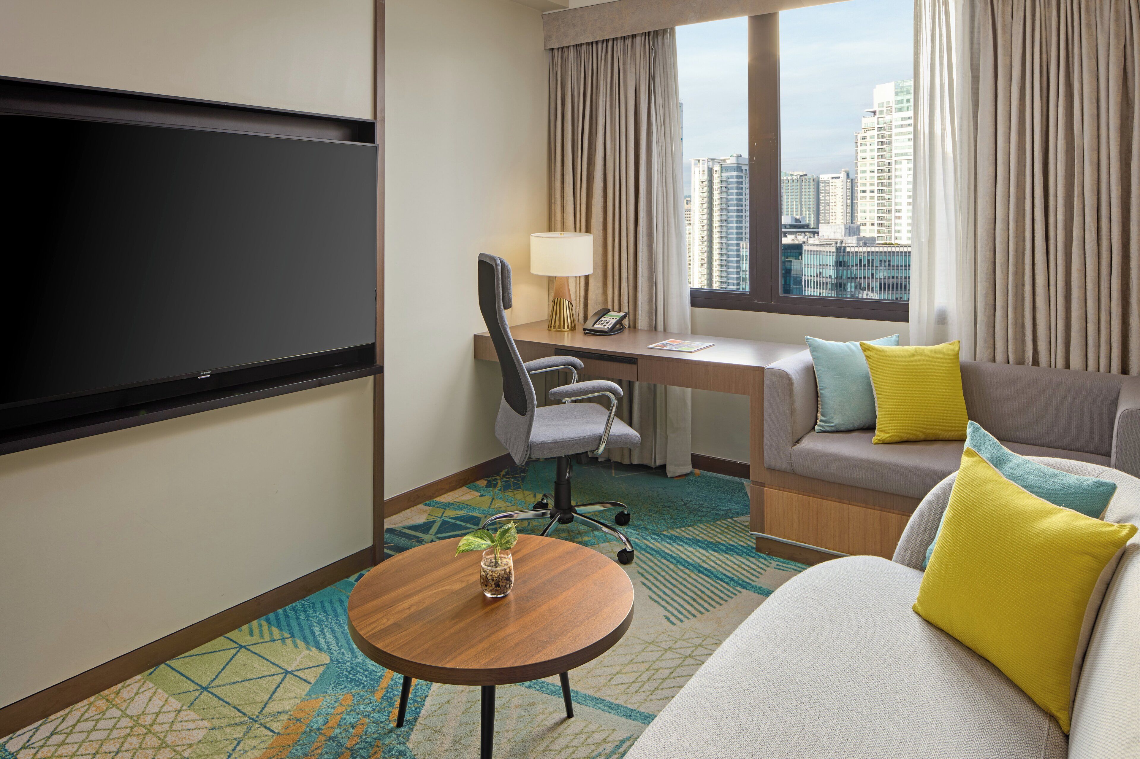 A hotel room with a bed, a desk, a chair, a TV, a lamp, a window with curtains, and a view of buildings.