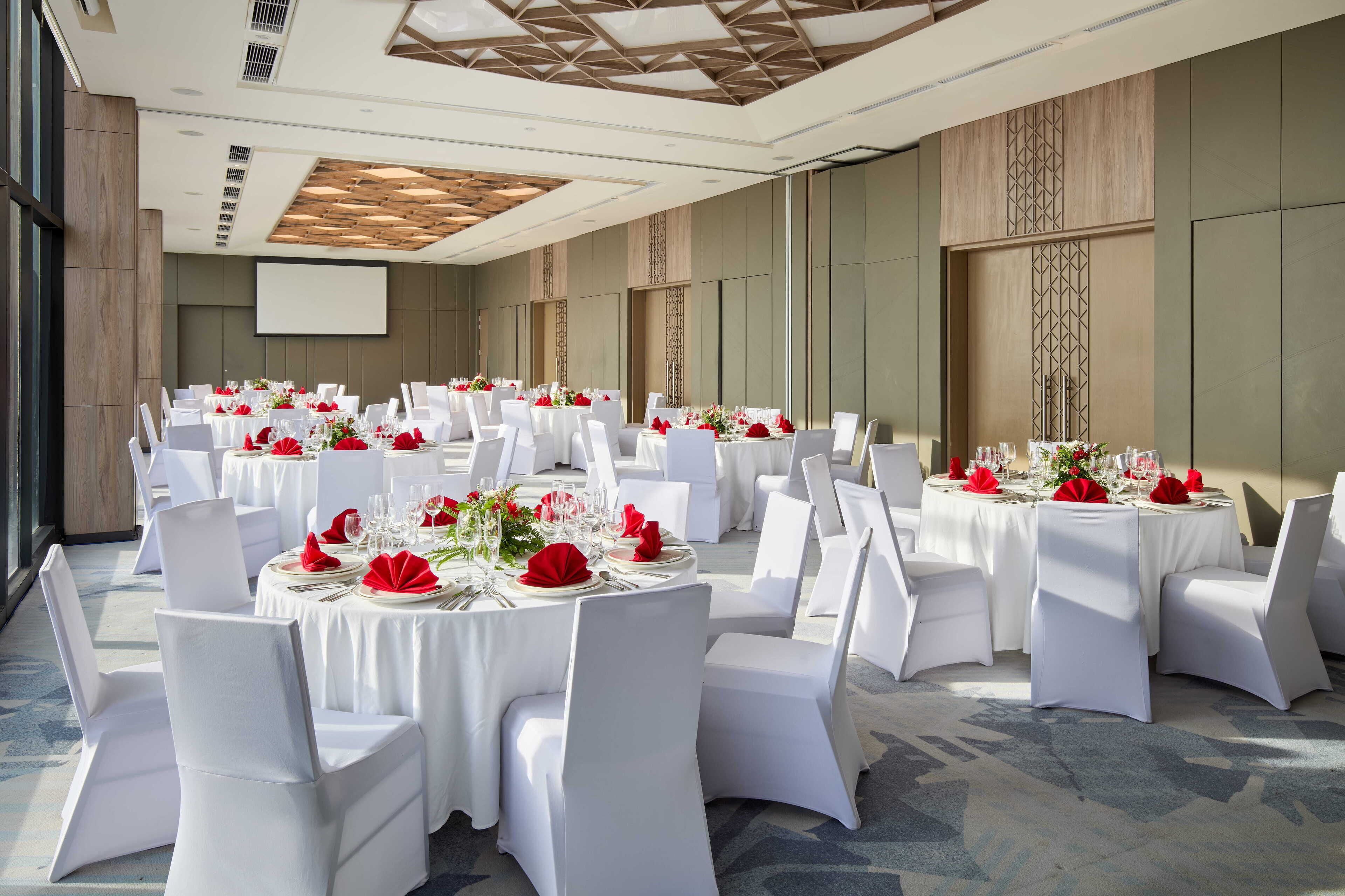 A banquet hall with round tables set for dining, white chairs, and red floral centerpieces.