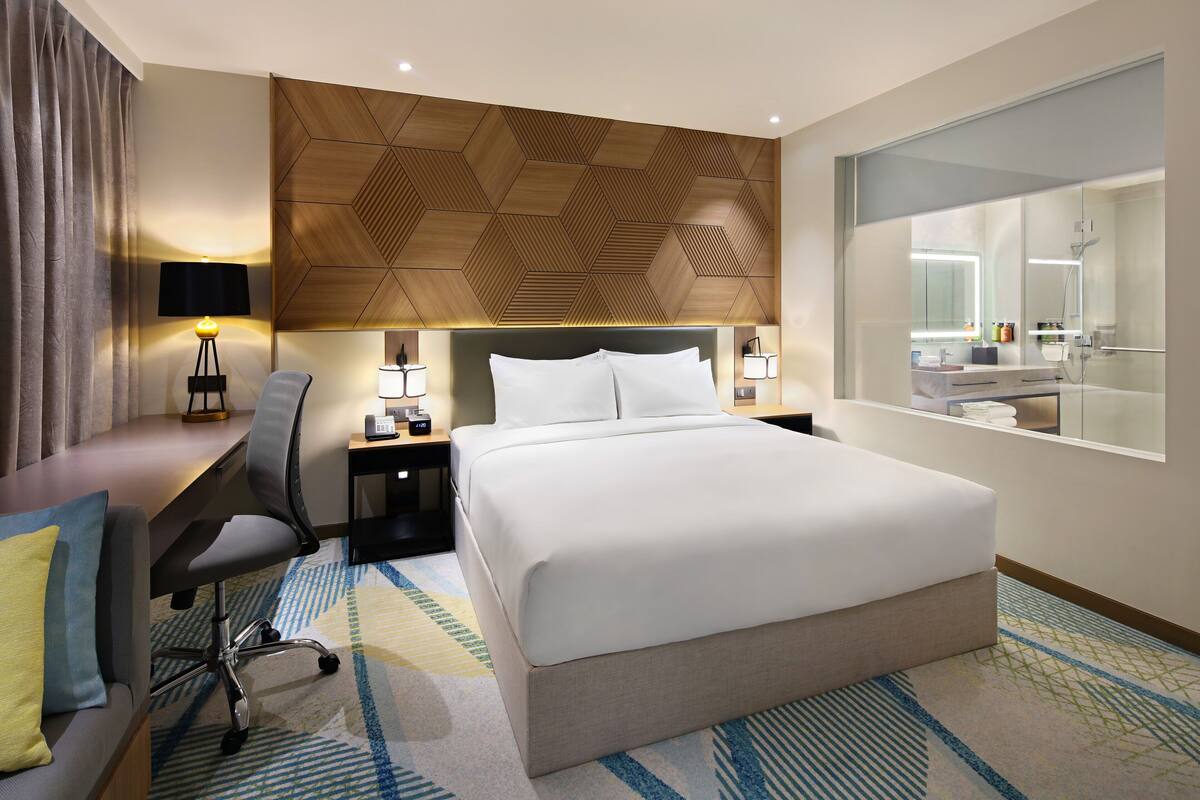 Une chambre d’hôtel moderne avec un grand lit, un bureau avec une chaise, une table de chevet, une lampe et un grand miroir.