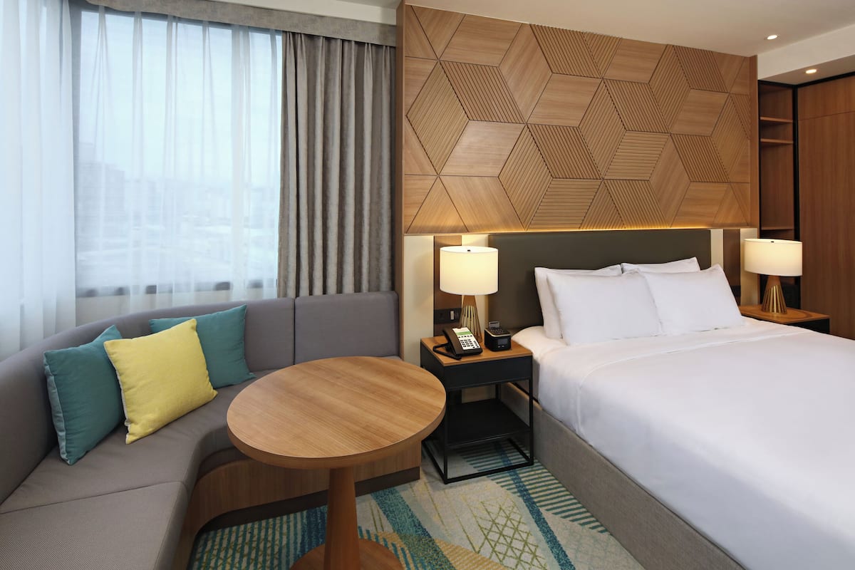 Une chambre d’hôtel comprenant un lit, un canapé, une table ronde, une table de chevet avec des lampes et une tête de lit ornée d’un motif géométrique.