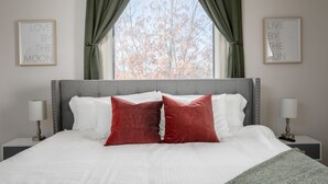 A neatly made bed with white and red pillows, a grey headboard, and two bedside tables with lamps. There are two framed quotes on the wall, one on each side of the window. - Mid-Century Modern Townhouse with High Views (Birmingham)