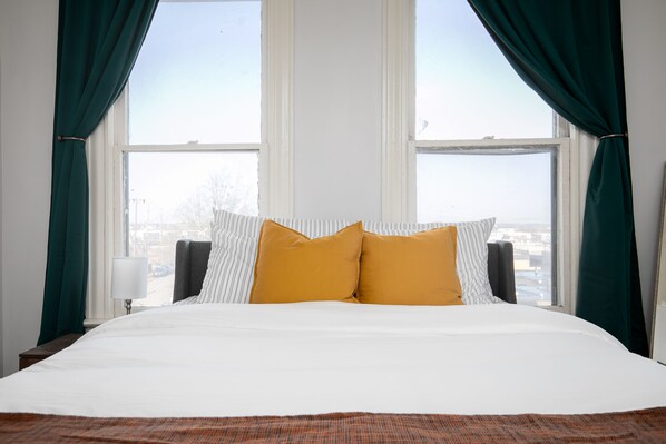 A hotel room with a large bed, two mustard-colored pillows, a striped pillow, and a view of the city through the window.