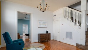 A living room with a glass coffee table, teal armchairs, a wooden cabinet, and a staircase with white railings. - Mid-Century Modern Townhouse with High Views (Birmingham)