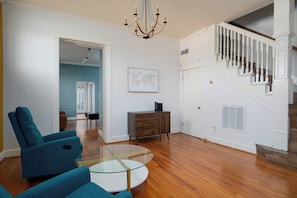 A living room with a glass coffee table, teal armchairs, a wooden cabinet, and a staircase with white railings.