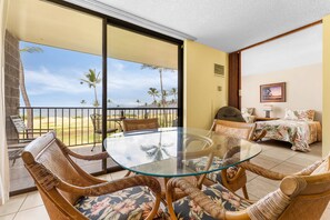 A room with a glass table and wicker chairs, a bed with floral bedding, and a balcony with a view of palm trees and the sea. - Kauhale Makai by Maui Rental Group (Kihei)