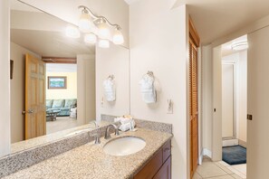 A bathroom with a granite countertop, a sink, a mirror, and a shower area. - Kauhale Makai by Maui Rental Group (Kihei)