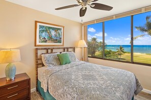A bedroom with a large window offering a view of the ocean, a ceiling fan, a bed with patterned bedding, and a nightstand with a lamp. - Kauhale Makai by Maui Rental Group (Kihei)