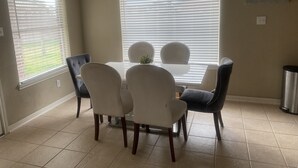 A dining area with a table and chairs, large windows with blinds, a ceiling light fixture, and a wall-mounted trash can.