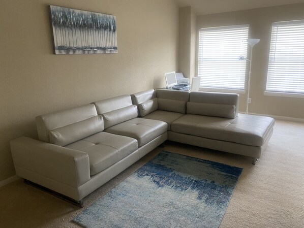 Un sofá seccional color beige en una sala de estar con un tapete azul y gris, un cuadro en la pared y una ventana con persianas.