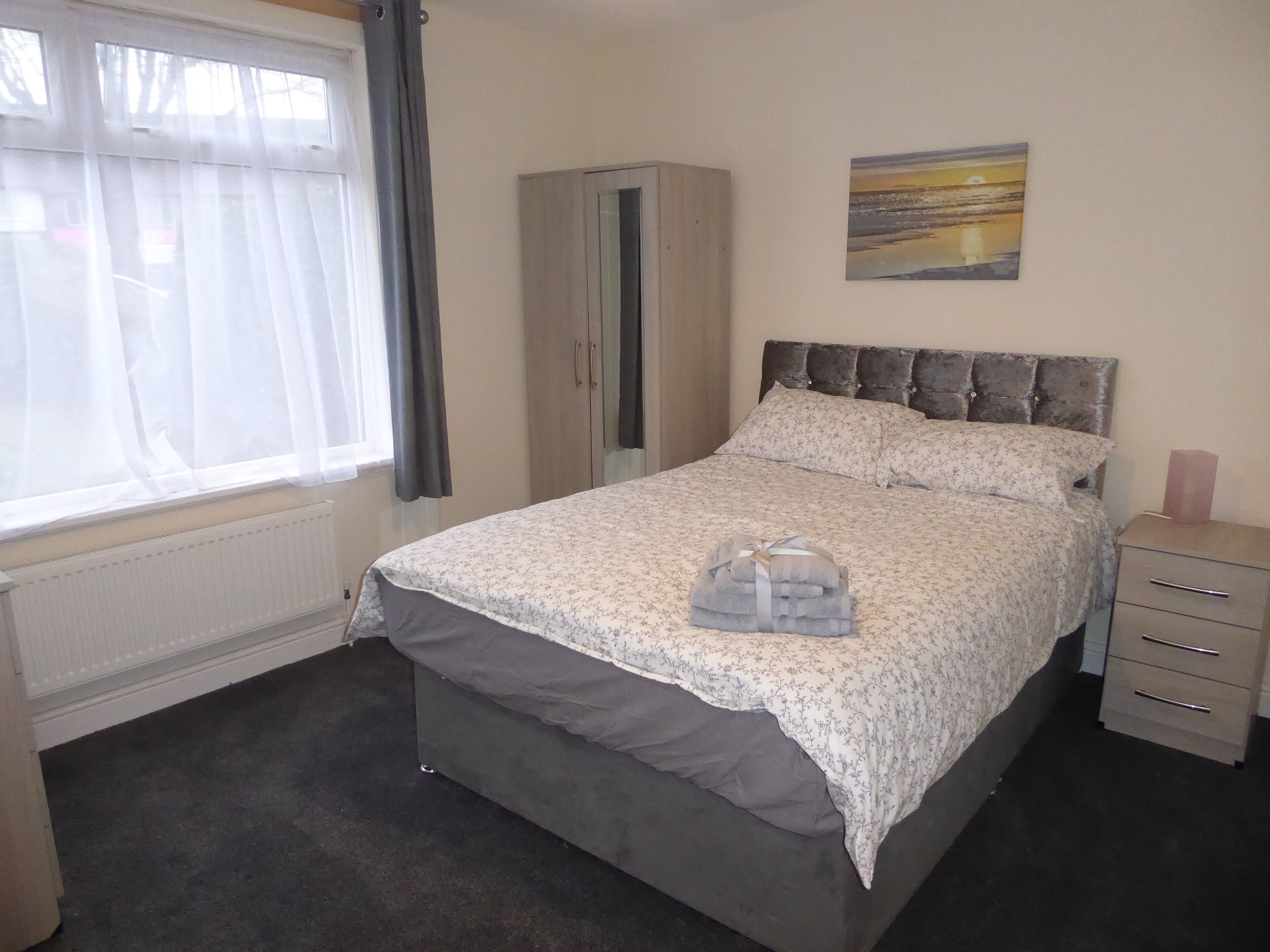 A bedroom with a bed, a wardrobe, a radiator, a window with curtains, and a painting on the wall.