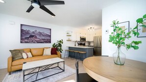 Una sala de estar moderna con sofá, una mesa de centro y una mesa de comedor. Hay una zona de cocina con refrigerador, microondas y taburetes de bar. Se ve un ventilador de techo, y una planta está colocada sobre la mesa de comedor.