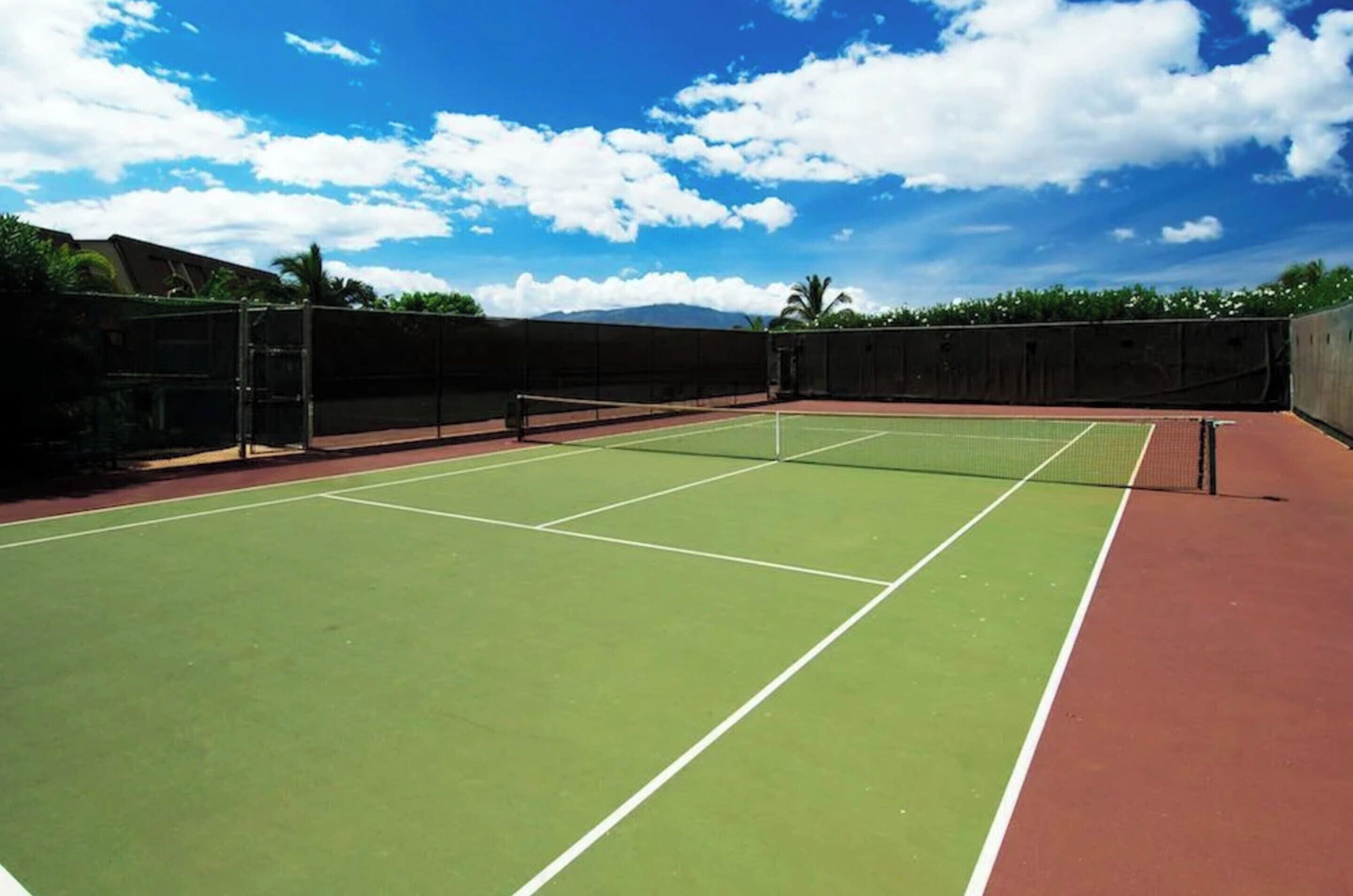 An outdoor tennis court with a net, surrounded by a fence and greenery.